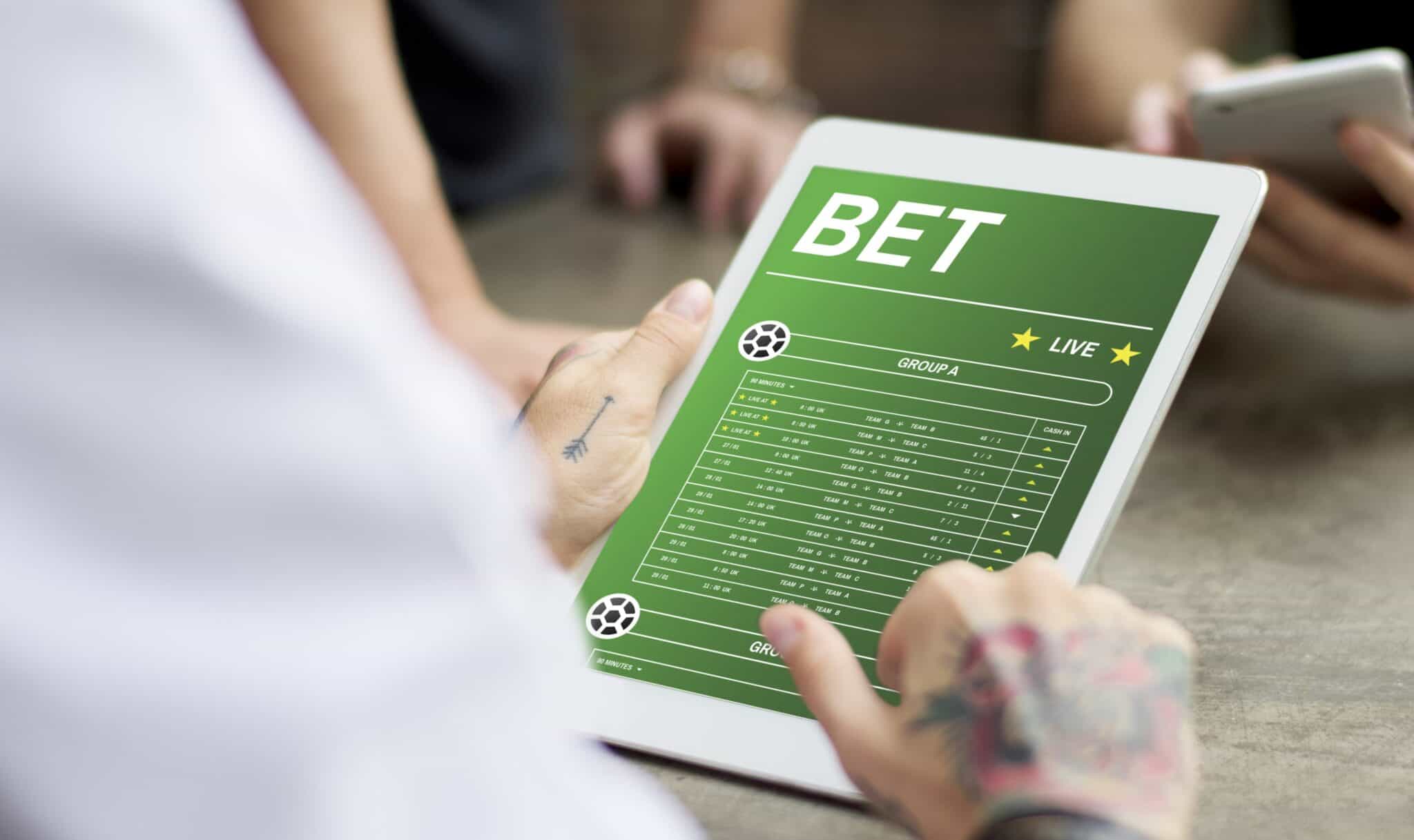 Imagem de um homem de costas segurando um tablet aberto em um site com informação de odds em apostas esportivas, exemplificando o que é e como funciona